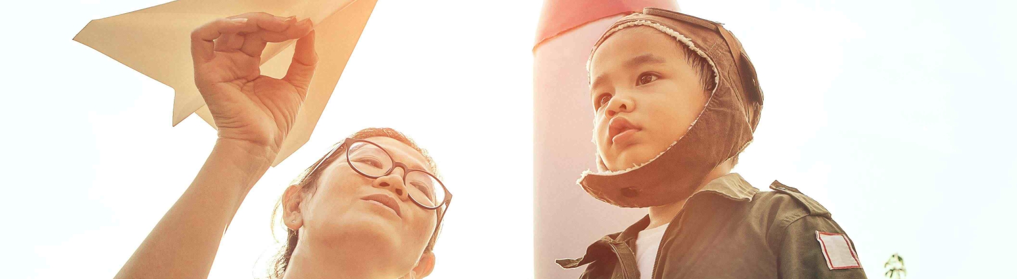 Picture of a woman holding a paper airplane and showing it to a little boy who is dressed like a pilot.